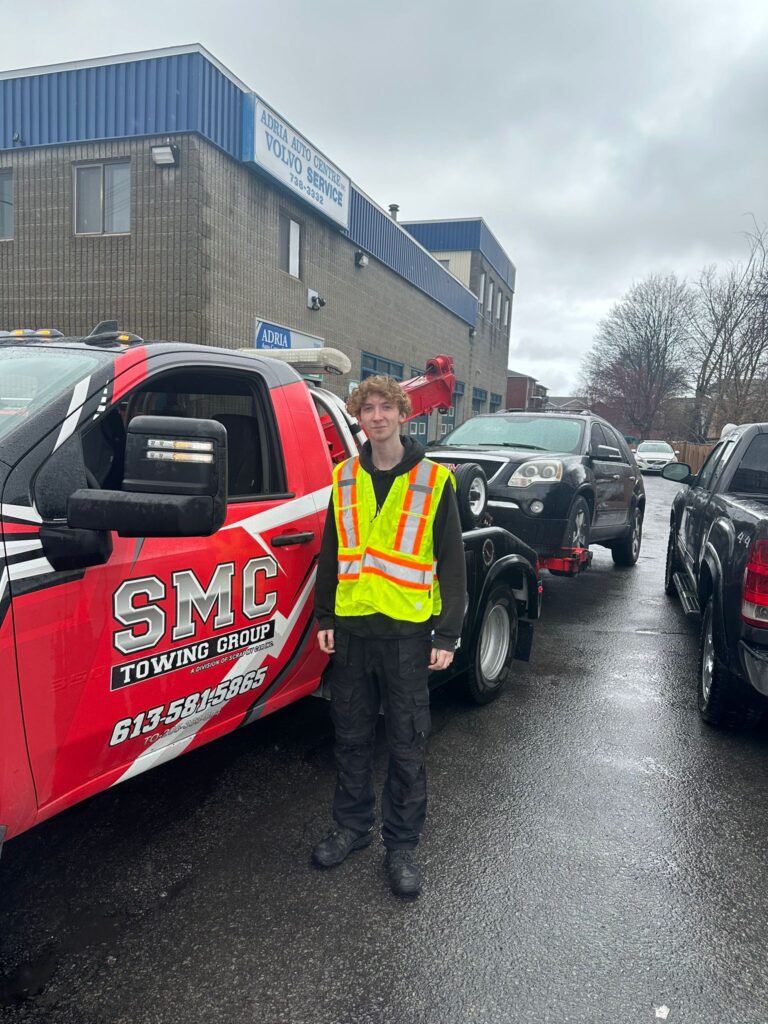 SMC Towing Group operator standing beside a tow truck with a vehicle loaded on a rainy day.